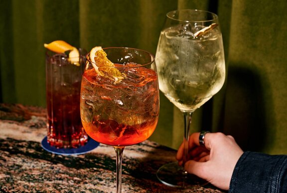 A person's hand, holding the stem of a white wine glass, two other cocktails next to it.