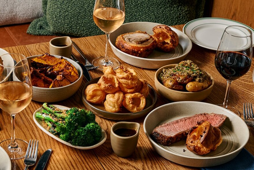 A table set with roast beef and pork, vegetables, yorkshire puddings and wine.