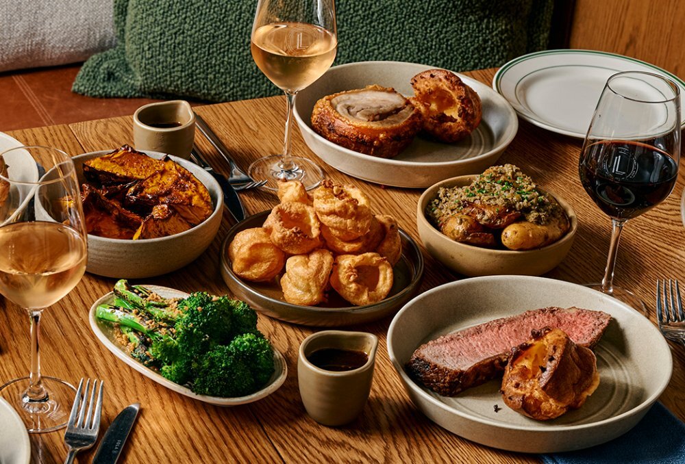 A table set with roast beef and pork, vegetables, yorkshire puddings and wine.