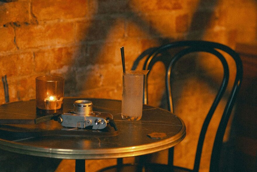 A small table for one with a sunset glow on the wall, a tall drink with a straw on the table and a votive candle.