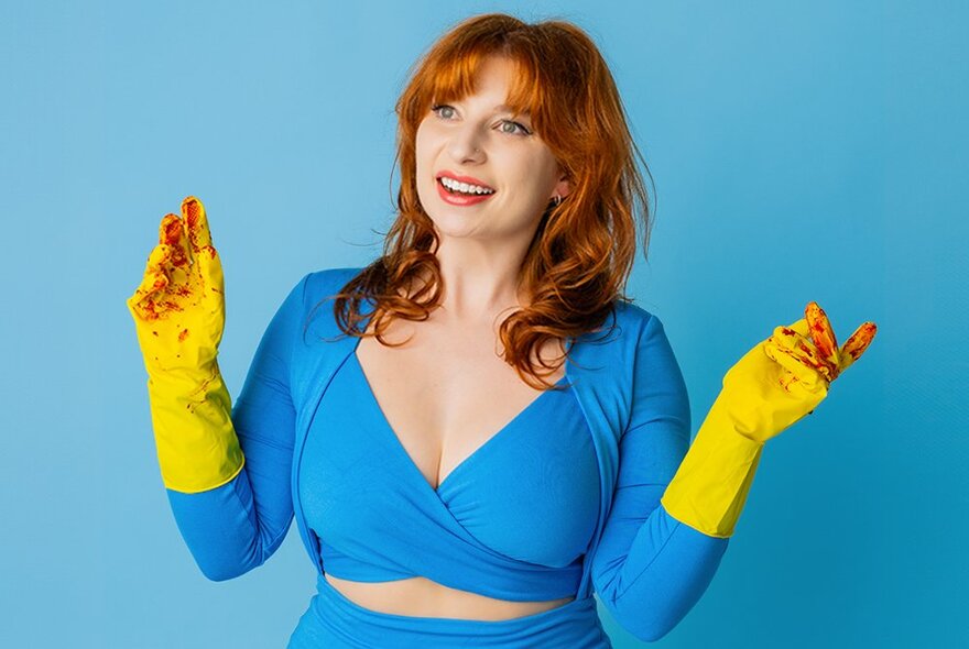 Performer Robyn Reynolds, a woman wearing an aqua coloured tight fitting dress, yellow rubbers gloves stained red at the fingertips, with shoulder length red hair and with a smile on her face.