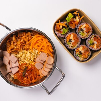 A Korean ramyun hot pot with sliced deli meats, sliced carrots, egg noodles and kimchi alongside a plate of Korean sushi rolls.