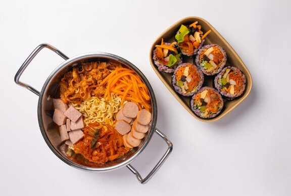 A Korean ramyun hot pot with sliced deli meats, sliced carrots, egg noodles and kimchi alongside a plate of Korean sushi rolls.