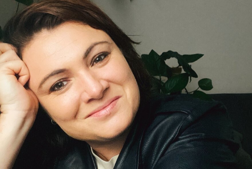 A smiling woman, resting her head on her propped up forearm, she has dark hair and there is a green plant behind her.