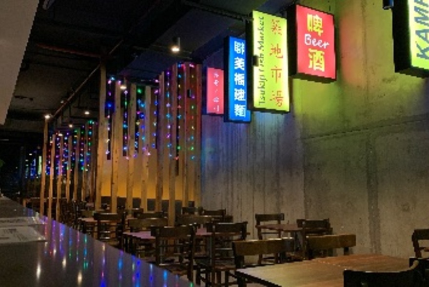 Interior of restaurant showing tables and chairs against a concrete wall, and illuminated signs hanging from the wall.