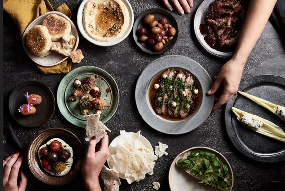 Hands dipping into multiple dishes of food served on a dark table including bread, dips and vegetables.