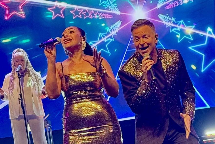 Singers Paulini and Tim Campbell on stage with a back-up singer and a projection of bright neon stars behind them.