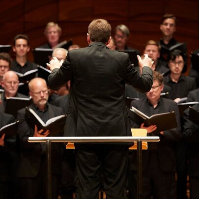 The back of a conductor as he conducts a choir in a concert hall. 