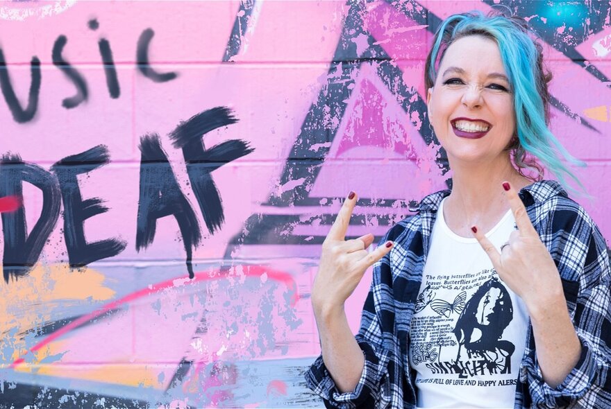 A woman with aqua blue hair wearing an open flannelette shirt in front of a pink graffitied wall with the word DEAF visible. 