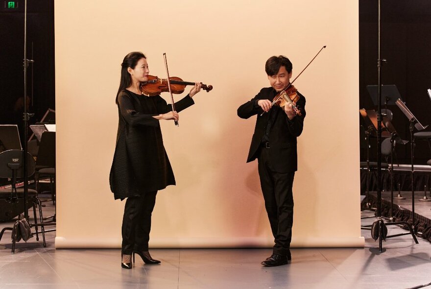 Two violinists stand playing in front of a cream-coloured backdrop.