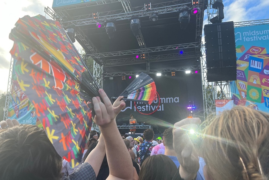 A large crowd at an outdoor stage, seen from eye level; people holding rainbow-coloured fans.