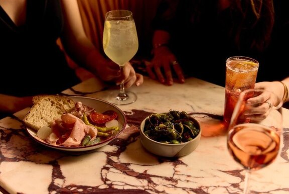 Two hands resting on a table in a restaurant, holding drinks, with small plates of charcuterie and a bowl of olives.