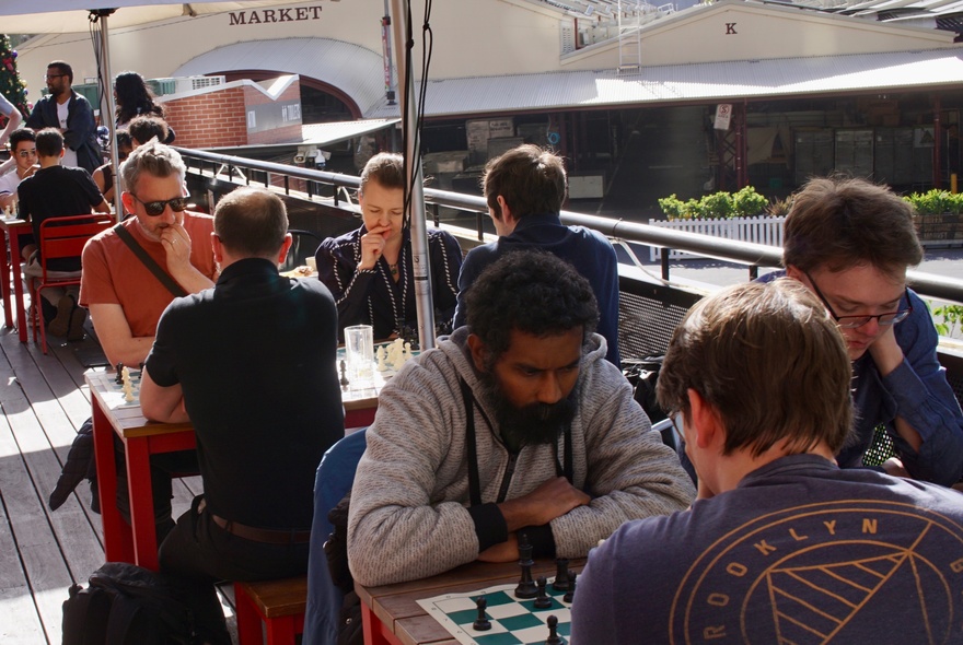 People playing chess at outdoor tables under unbrellas on a raised terrace.