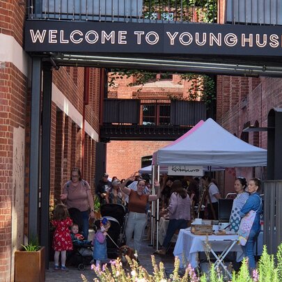 Market stall tents with signage next to a heritage brick building, with greenery and families.
