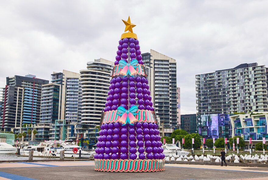 A giant Christmas tree with purple baubles and a gold star, with city buildings in the background.