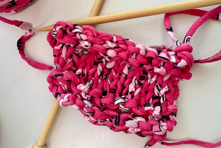 A knitting project being worked on with bright pink and variegated t-shirt yarn using wooden knitting needles.