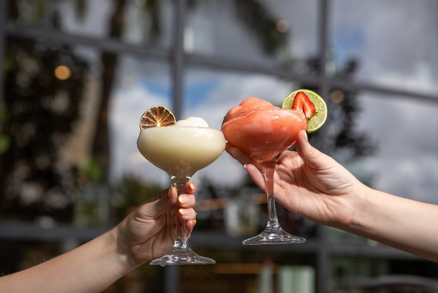 Two hands coming together holding glasses of a coloured cocktail and clinking them together in a celebratory way.