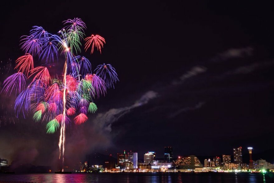 City skyline at night with bright purple, red and green fireworks visible in the distance.