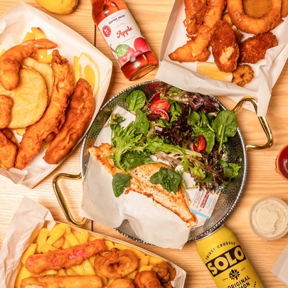 Looking directly down on a table with boxes of fish and chips, a plate with grilled fish and salad, various soft drinks and condiments. 