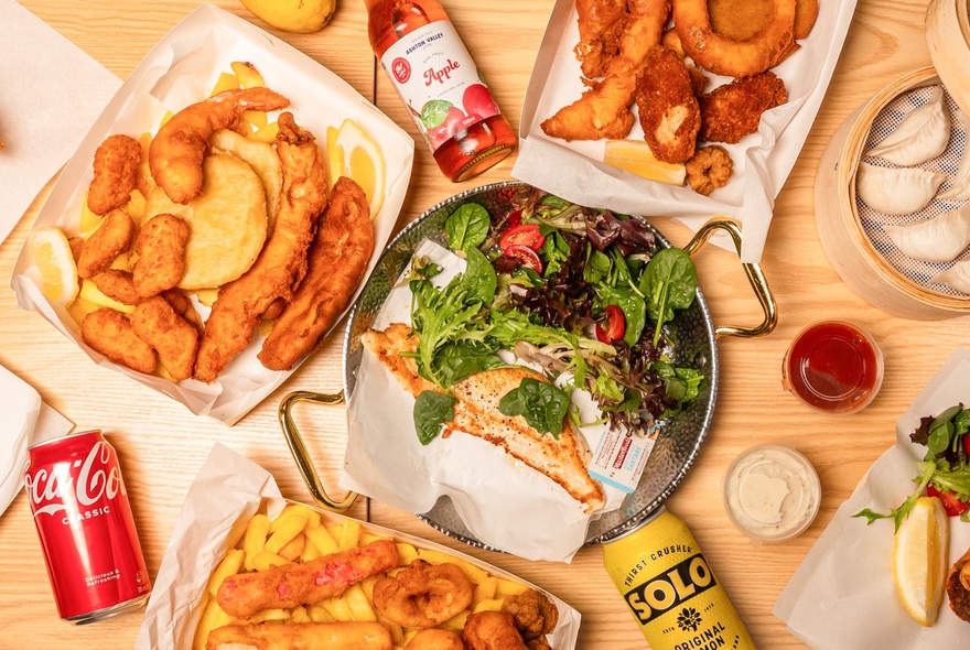 Looking directly down on a table with boxes of fish and chips, a plate with grilled fish and salad, various soft drinks and condiments. 