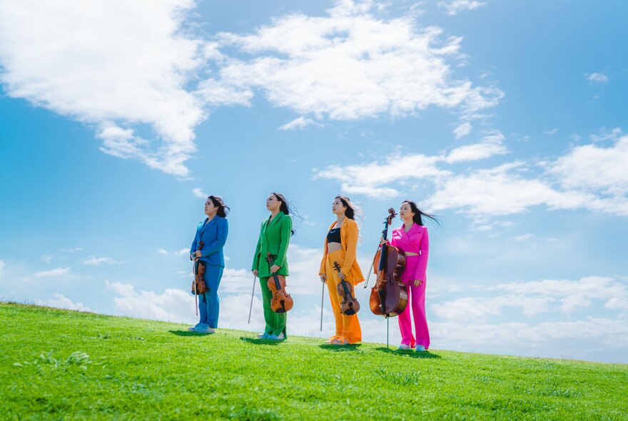 The four members of Invictus Quartet, each wearing a different coloured suit, standing in a line on a grassy hill against a blue sky background.