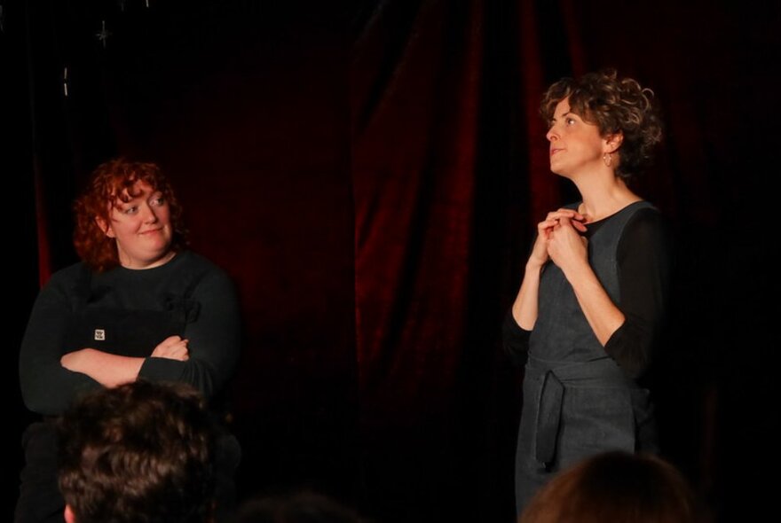 Two actors improvising on a theatre stage in front of a black curtain, a live audience watching.