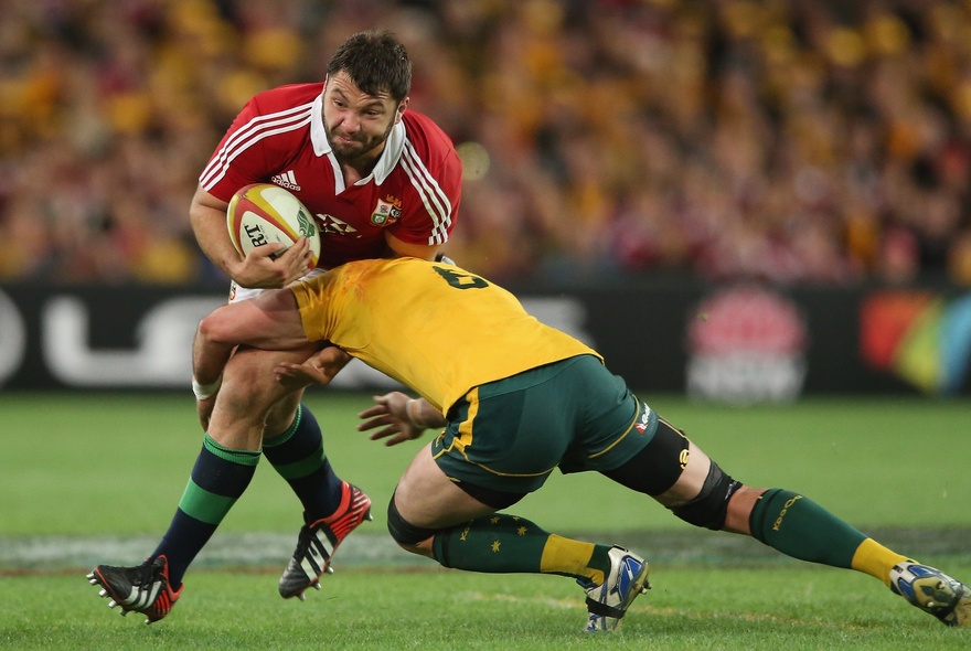 One rugby player tackling another rugby player who is holding the ball, on a green sporting field. 