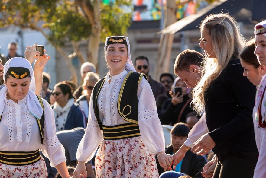 Women on traditional costumes assembled for a dance in a crowded area outdoors. 