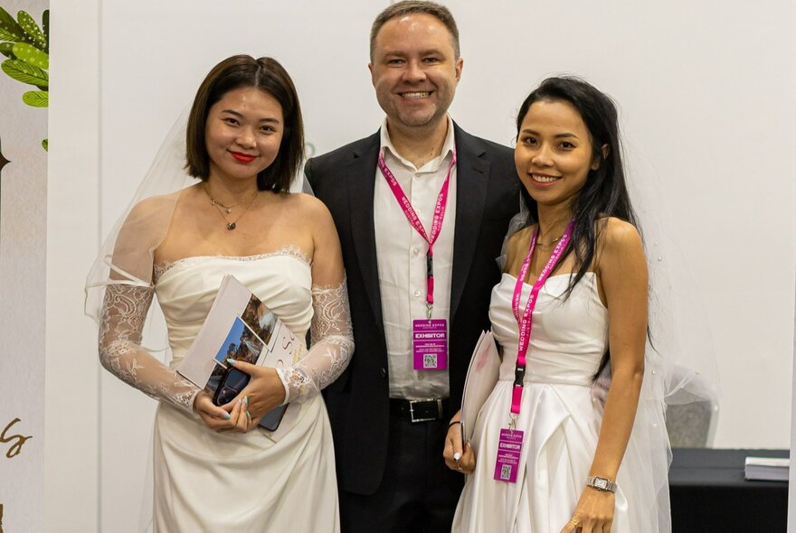 Two women in white bridal dresses next to a smiling man at an expo.