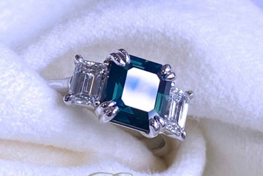 A white gold engagement ring with a central teal blue sapphire flanked by two emerald cut diamonds, nestled in a soft, cream-coloured cloth.