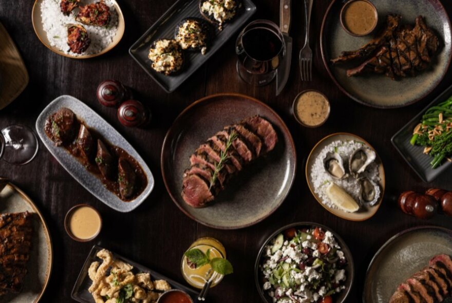 Looking down on a table of many dishes, the centrepiece being a plate of sliced steak. 