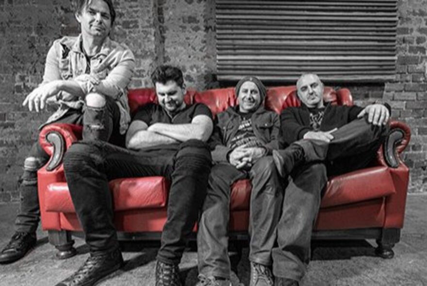 Four men slouching on a red couch.