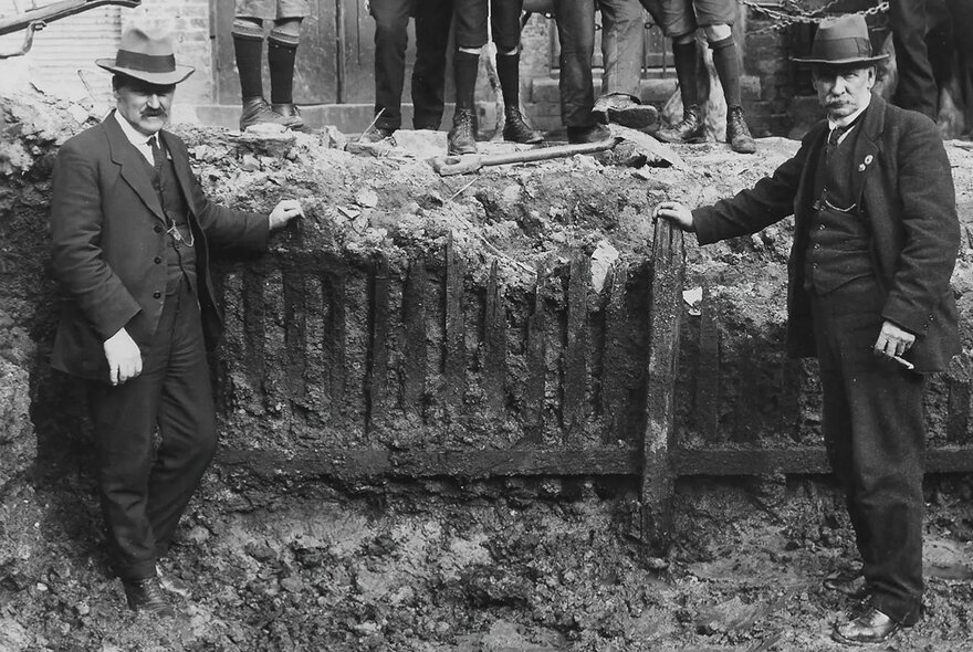 Two men in suits and hats dating to the mid-1850s standing in a muddy hole, with dirt and a fence between them, and the boots of men holding spades in the background behind them.