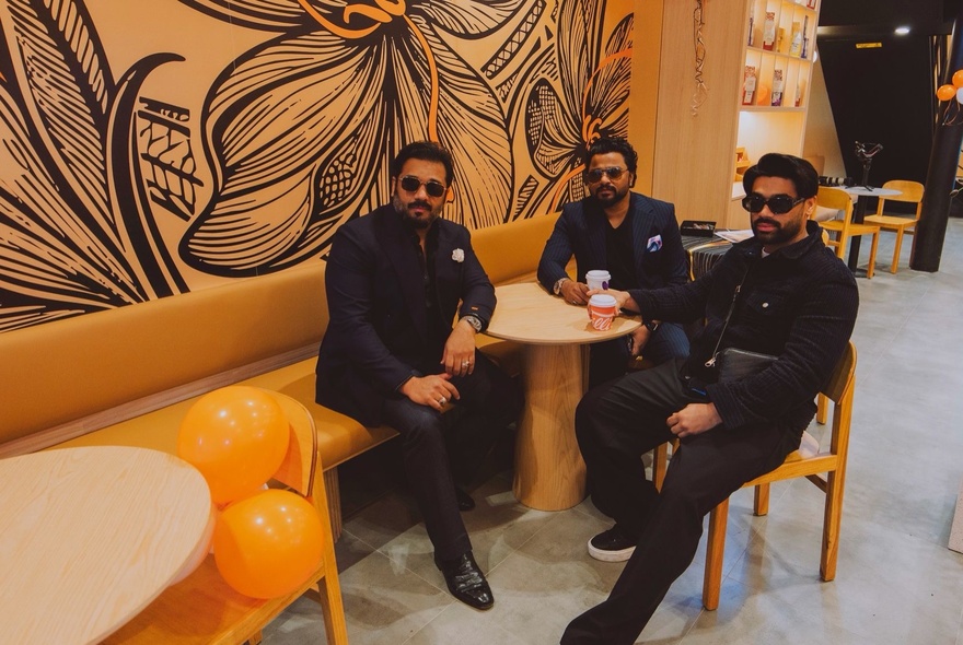 Three men in dark suits wearing sunglasses, sitting at a small table inside an orange-themed cafe with a flower mural on the wall behind them.