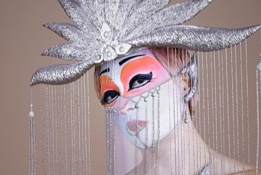 A non-binary individual with traditional Asian-inspired full face paint behind a shimmering curtain of silver strings falling from a silver headdress.