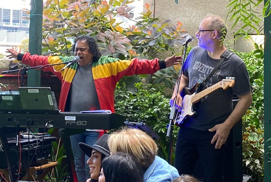Soul musician Dinash Daniel, standing on stage behind his keyboard, arms outstretched, the guitar player looking over at him.