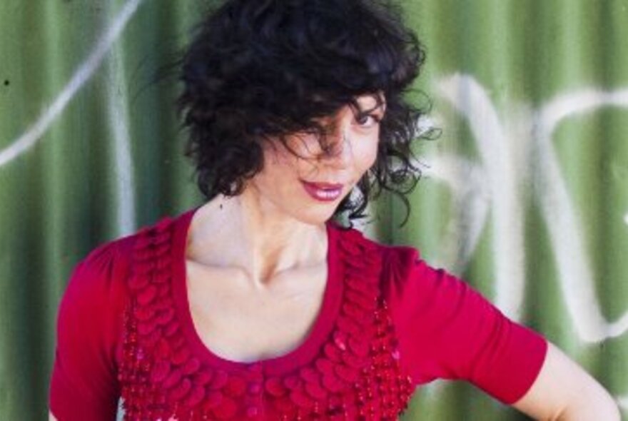 Woman with dark hair, partly over face, smiling wearing a crimson dress, standing against a graffitied green corrugated-iron wall.