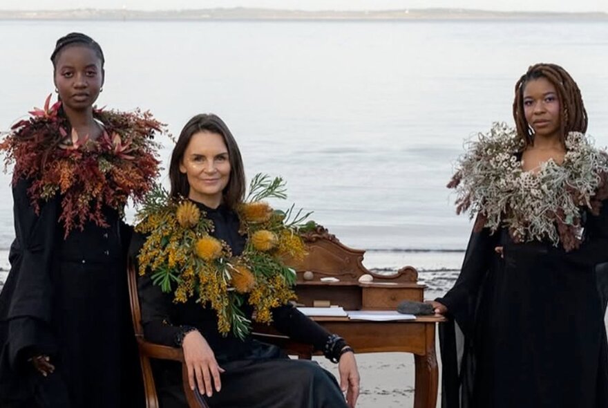 Three women on a beach on a grey day, one seated at an antique writing desk, each wearing a black robe with a wreath of Australian native flowers around their necks.