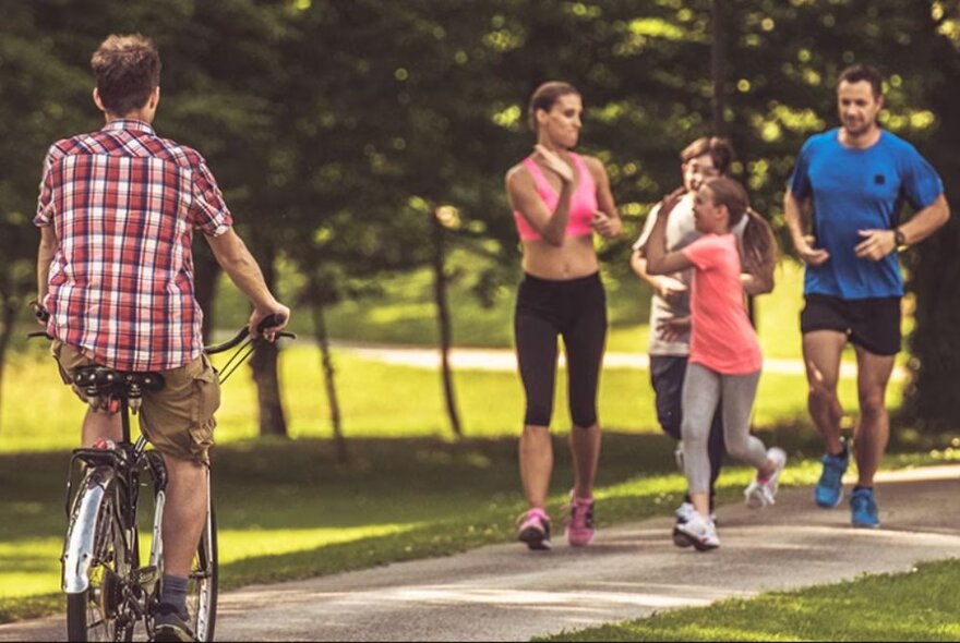 A family running and a man riding a bike along a path with grass and trees in the background.