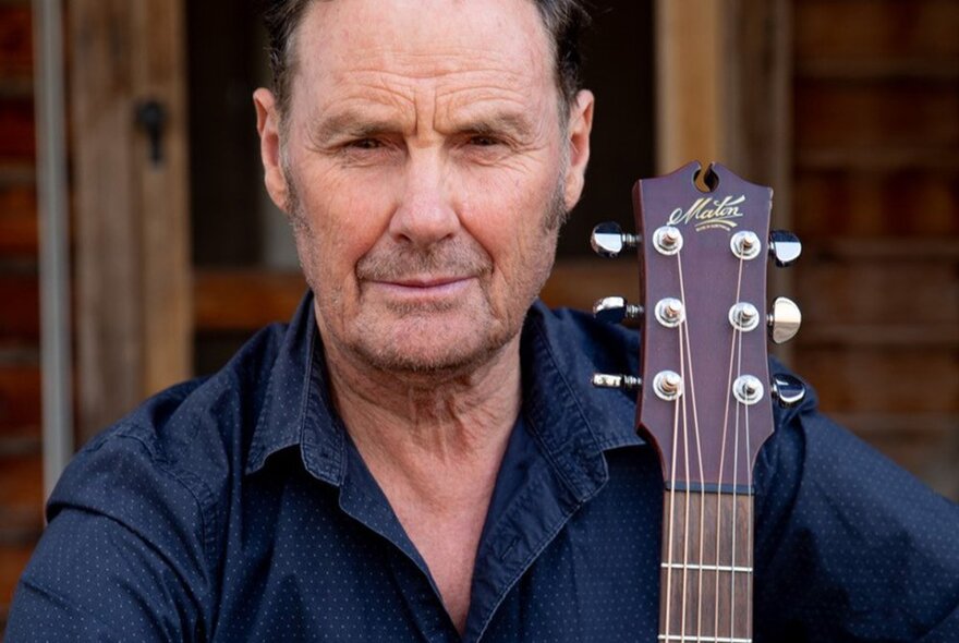 Portrait of Neil Murray in front of a wooden homestead, wearing a dark blue shirt and holding his acoustic guitar.