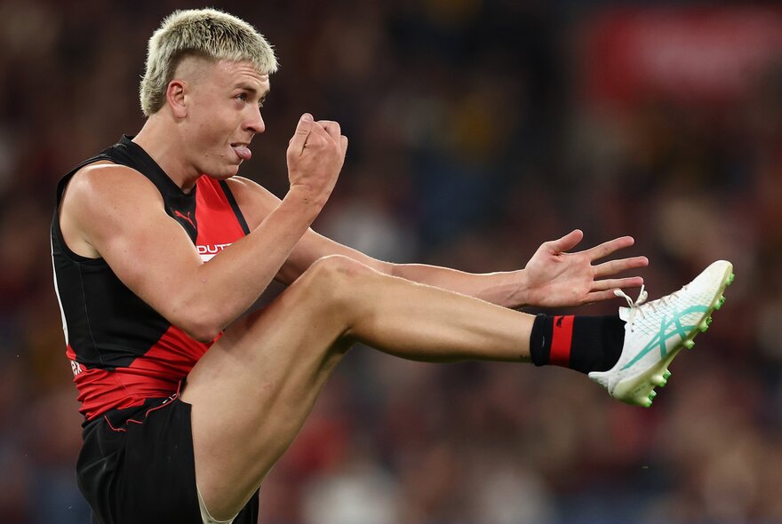 Essendon AFL player Nate Caddy after kicking a football, lef in the air and tongue out. 