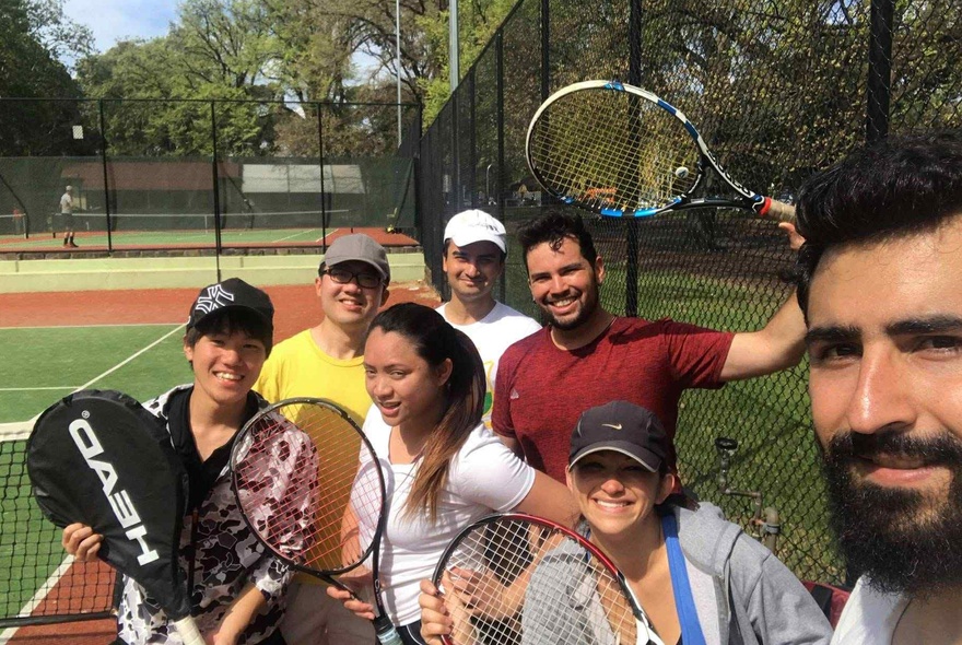 A group of smiling people standing on a tennis court, some holding racquets, taking a selfie