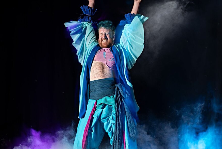 A man on stage with his arms in the air, wearing a vibrant blue ruffled outfit with a teal cummerbund.