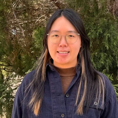 A person wearing glasses and a dark buttoned shirt standing outdoors in front of leafy foliage.