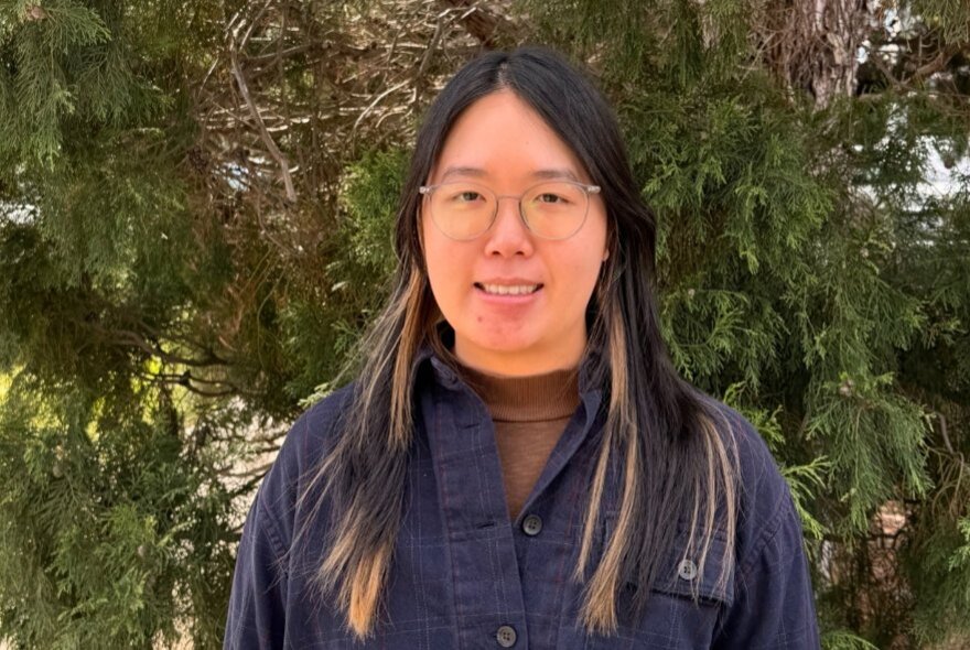 A person wearing glasses and a dark buttoned shirt standing outdoors in front of leafy foliage.