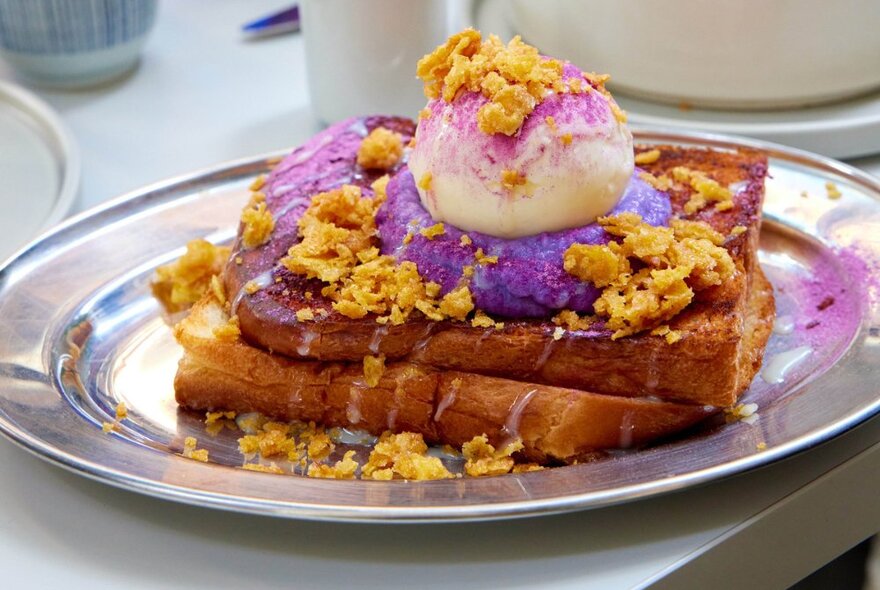 Thick piece of brioche toast on a silver plate topped with ube, ice cream and cornflakes.