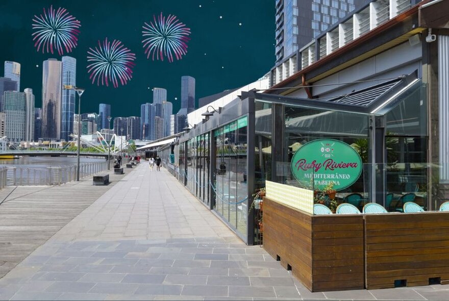 The waterfront and paved wharf area in front of Ruby Riviera restaurant, with an illustration of fireworks in the background over the top of city buildings.