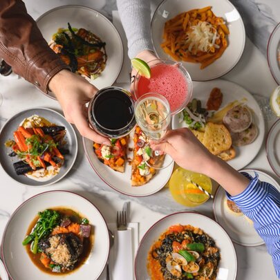 Looking down at multiple plates of Italian food on a table covered in white linen, with three hands clinking drinks in glasses over the food.