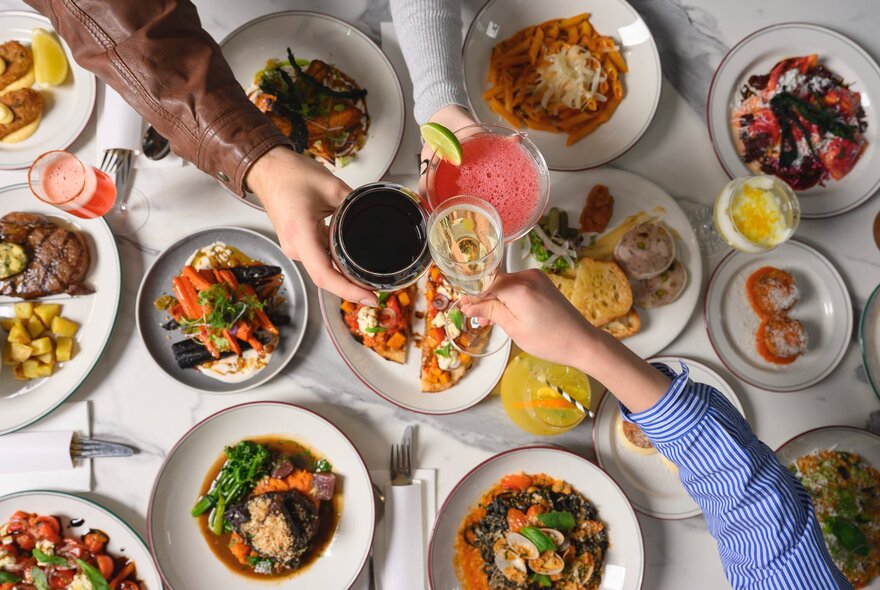 Looking down at multiple plates of Italian food on a table covered in white linen, with three hands clinking drinks in glasses over the food.