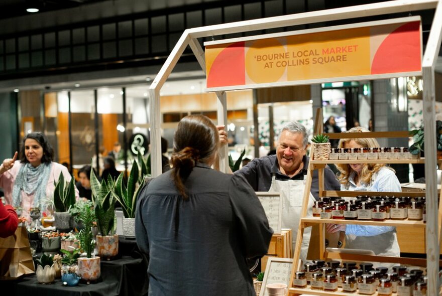 Two people interacting over a market stall selling produce in jars inside a retail shopping plaza.